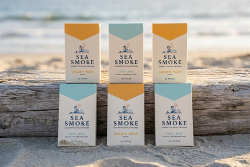 Six Sea Smoke packs in two rows on a driftwood log, three Golden Hour and three Surf Wax